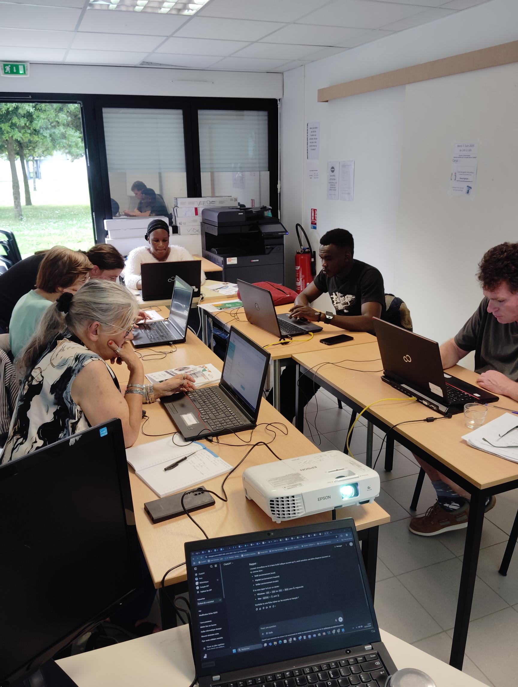Photo d'un atelier d'aide numérique se déroulant dans la salle d'atelier de la Conciergerie 5 personnes assises, chacune derrière un ordinateur portable et une personne debout, penchée sur l'écran d'une des personnes assise, pour aider cette dernière . Cette photo est prise dans la salle d'atelier de la Conciergerie AcSoMur où se déroule les ateliers d'aide numérique