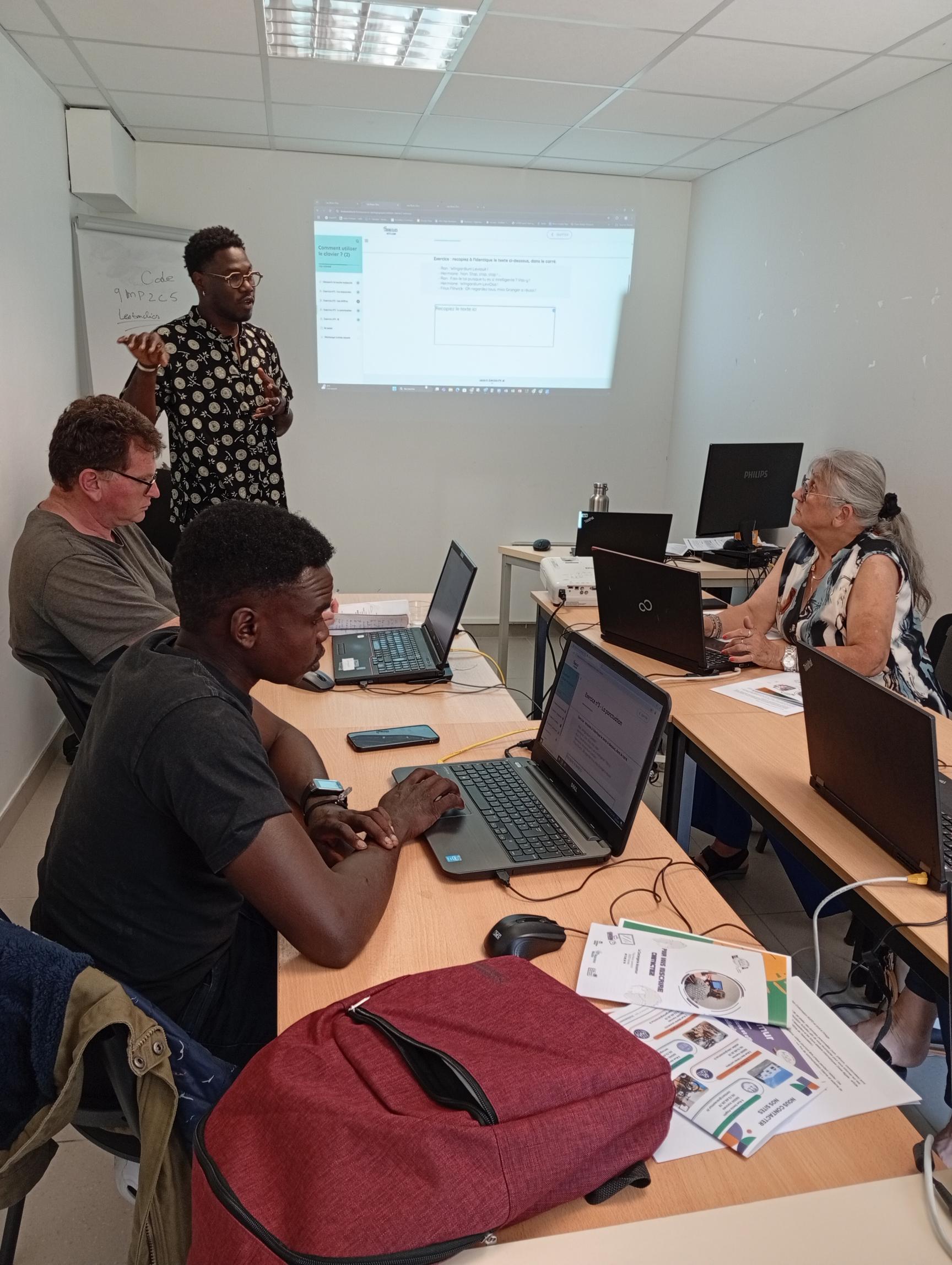Photo d'un atelier d'aide numérique se déroulant dans la salle d'atelier de la Conciergerie  3 personnes assises, chacune derrière un ordinateur portable et une personne debout devant un éctan projeté. Cette photo est prise dans la salle d'atelier de la Conciergerie AcSoMur où se déroule les ateliers d'aide numérique